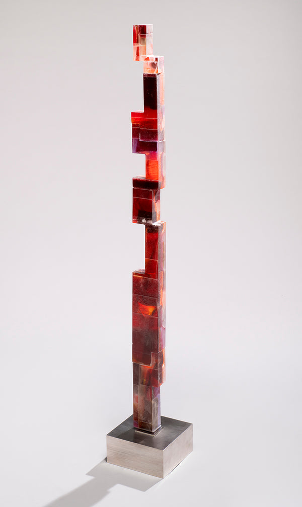 Tall glass sculpture of stacked, offset blocks in deep red and amber rising from a brushed stainless steel base.