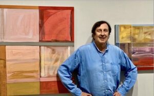 A photo of artist David Kupferman standing in front of two geometric paintings